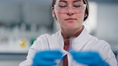 Young adult woman wearing safety glasses and lab coat, holding test tube while performing science research in a chemistry lab