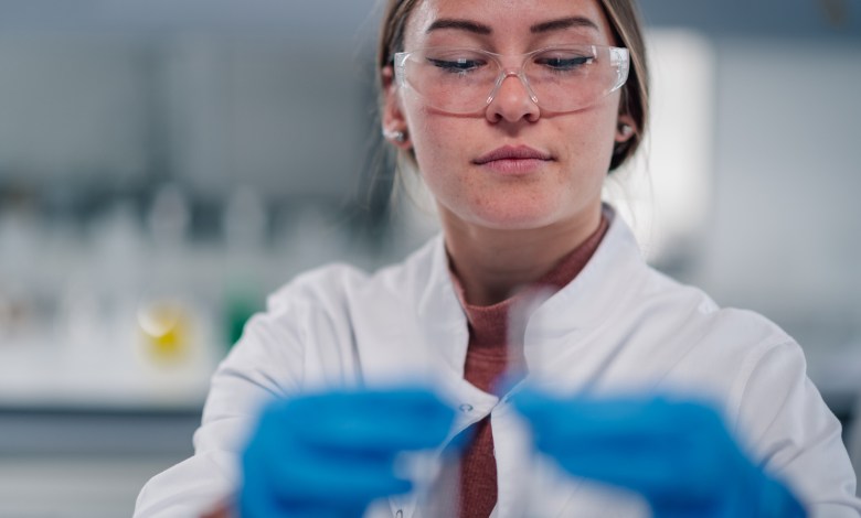 Young adult woman wearing safety glasses and lab coat, holding test tube while performing science research in a chemistry lab
