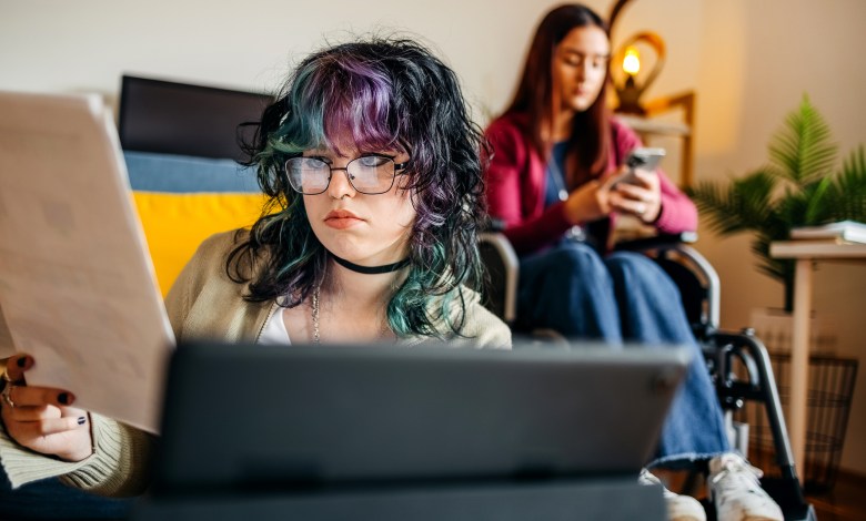 Young female adult with disability preparing for exam studying together with friend at home who is looking at charts while sitting on floor in front of digital tablet