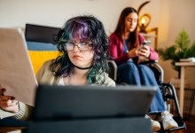Young female adult with disability preparing for exam studying together with friend at home who is looking at charts while sitting on floor in front of digital tablet