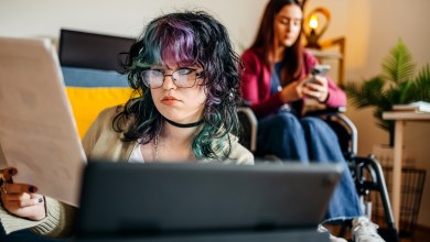 Young female adult with disability preparing for exam studying together with friend at home who is looking at charts while sitting on floor in front of digital tablet