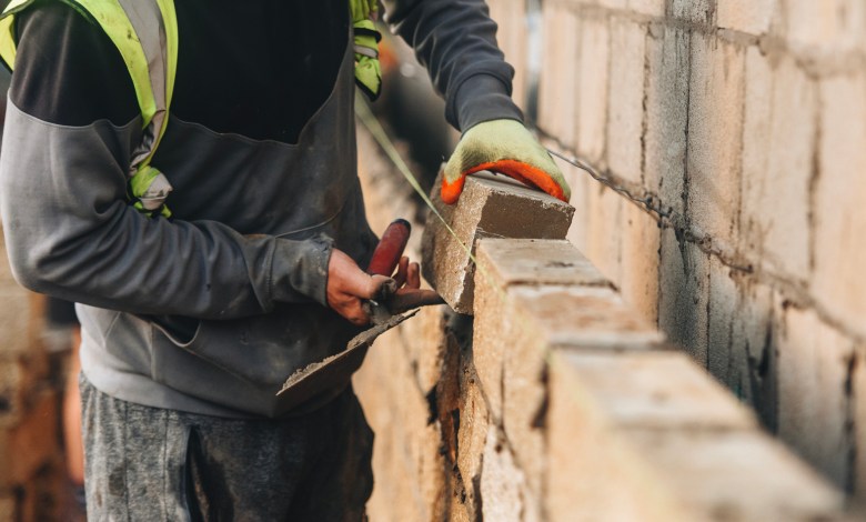 Bricklayer constructing stone wall using stone blocks, cement mortar mix and trowel