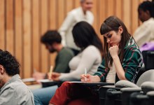 Students sitting in a modern lecture hall, writing papers and learning. An instructor is walking through the aisles